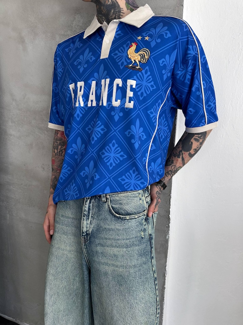 Peut inclure: Un polo bleu &agrave; manches courtes avec un col blanc, des passepoils blancs et un motif r&eacute;p&eacute;t&eacute; de fleurs de lys. Le mot FRANCE est imprim&eacute; en blanc sur la poitrine, accompagn&eacute; d'un embl&egrave;me de coq dor&eacute; et de deux petites &eacute;toiles dor&eacute;es au-dessus.