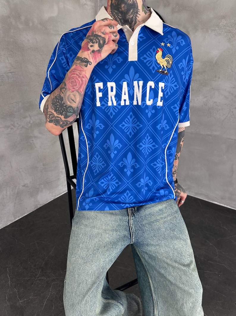 Peut inclure: Un polo bleu avec un col blanc, des passepoils blancs et un motif r&eacute;p&eacute;titif de fleurs de lys. Le mot FRANCE est imprim&eacute; en blanc sur la poitrine, accompagn&eacute; d'un embl&egrave;me de coq dor&eacute; avec deux &eacute;toiles au-dessus sur le c&ocirc;t&eacute; gauche.