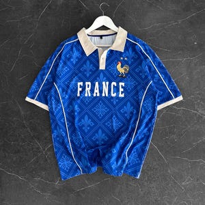 Peut inclure: Un polo bleu &agrave; manches courtes avec un motif r&eacute;p&eacute;t&eacute; de fleurs de lys, un col beige et un passepoil blanc. Le mot FRANCE est imprim&eacute; en blanc sur la poitrine, accompagn&eacute; d'un graphique repr&eacute;sentant un coq et deux &eacute;toiles au-dessus.
