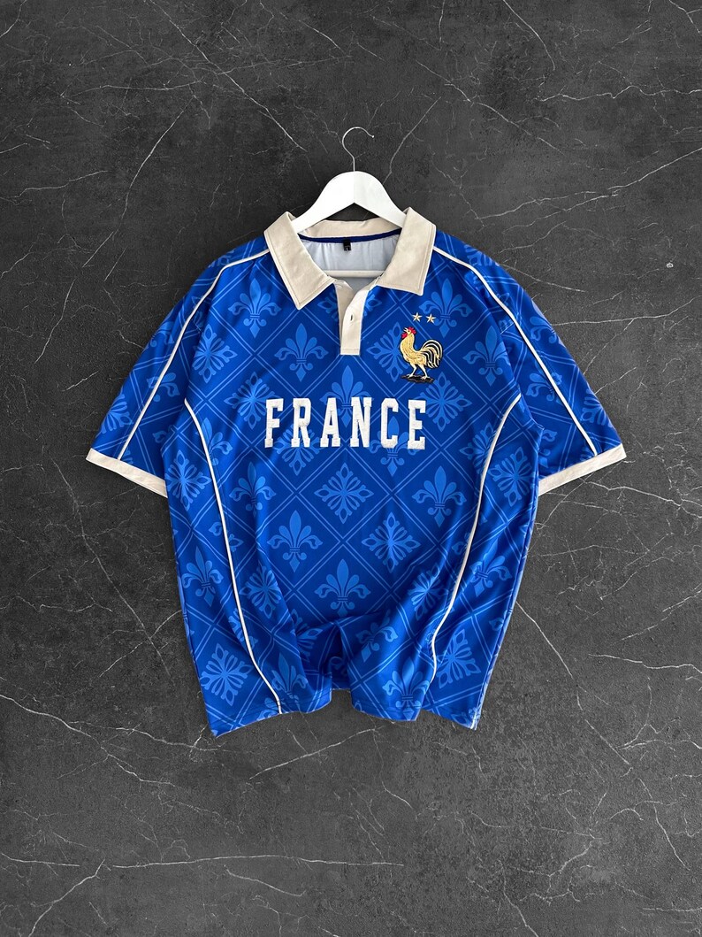 Peut inclure: Un polo bleu &agrave; manches courtes avec un motif r&eacute;p&eacute;t&eacute; de fleurs de lys, un col beige et un passepoil blanc. Le mot FRANCE est imprim&eacute; en blanc sur la poitrine, accompagn&eacute; d'un graphique repr&eacute;sentant un coq et deux &eacute;toiles au-dessus.