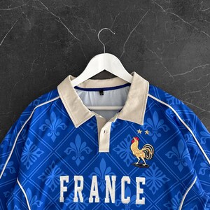 Peut inclure: Un polo bleu avec un motif r&eacute;p&eacute;titif de fleurs de lys bleu clair dans un quadrillage. Le v&ecirc;tement pr&eacute;sente le mot FRANCE en lettres capitales blanches, un embl&egrave;me de coq dor&eacute; surmont&eacute; de deux &eacute;toiles jaunes et un col beige.