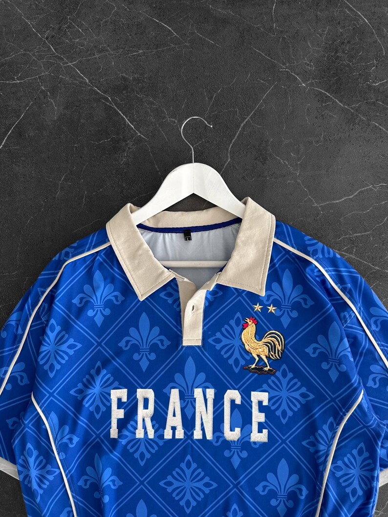Peut inclure: Un polo bleu avec un motif r&eacute;p&eacute;titif de fleurs de lys bleu clair dans un quadrillage. Le v&ecirc;tement pr&eacute;sente le mot FRANCE en lettres capitales blanches, un embl&egrave;me de coq dor&eacute; surmont&eacute; de deux &eacute;toiles jaunes et un col beige.