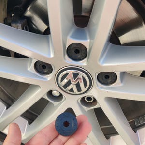 Könnte beinhalten: Eine dunkelgraue, kreisförmige Volkswagen Radschraubenabdeckung mit sternförmigem Mittelloch und dem eingeprägten Wort VOLKSWAGEN am Rand, gehalten vor einem silbernen Mehrspeichen-Autofelgenrad mit passenden installierten Abdeckungen und Logo.