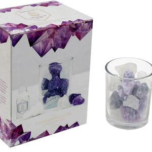 May include: A white box with purple crystal designs, labelled "Amethyst, The Stone of Peace", next to a glass jar filled with amethyst and clear quartz crystals. A small glass bottle with a dropper is also visible.