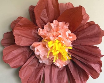 May include: A large, handmade paper flower with layered petals in shades of dusty rose and coral. The center features a cluster of peach-colored paper with yellow fringe, creating a vibrant and textured floral decoration.