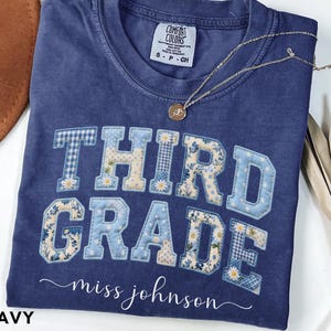 May include: A navy blue short-sleeve t-shirt featuring the text THIRD GRADE in large, block-style letters with a light blue and white floral and gingham patchwork pattern. Below the text, the name miss johnson is printed in white cursive script.