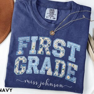 May include: A navy blue short-sleeve t-shirt with a crew neckline. The front features the text FIRST GRADE in block letters with a blue and white floral and gingham patchwork pattern. Below, the text miss johnson is printed in white script.