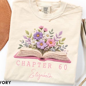 May include: An ivory t-shirt featuring a graphic of an open book with a bouquet of purple, pink, and white flowers emerging from the pages. Below the book, the text reads "CHAPTER 60" in a bold font and "Elizabeth" in a script font.