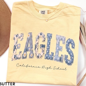 May include: A butter yellow crewneck t-shirt featuring the word EAGLES in large, patchwork-style letters with various blue and white floral, gingham, and polka dot patterns. Below, the text California High School is printed in black script.