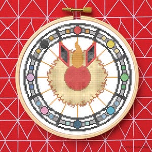 Puede incluir: Un diseño de punto de cruz circular terminado en un bastidor de madera. El diseño presenta un motivo central rojo y amarillo, rodeado por un borde circular con acentos coloridos. El fondo es una tela roja con un patrón geométrico blanco.