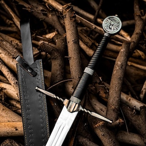 May include: A steel sword with a black wrapped handle and a circular pommel featuring a wolf head emblem. The sword includes a matching black leather scabbard with decorative stitching and a long strap, showcasing a metallic crossguard design.