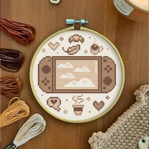 May include: A cross-stitch design in a gold hoop features a video game console with a cloudscape on the screen. The design includes hearts, coffee cups, and other game-themed elements in shades of brown and beige.