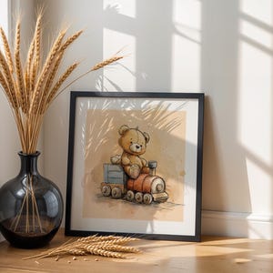 May include: A framed watercolour print depicts a brown teddy bear on a red and grey toy train. The artwork has a beige background with watercolour effects. The print is displayed on a wooden surface.
