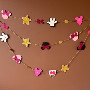 Puede incluir: Guirnalda decorativa con formas variadas: orejas de Mickey, lazos, estrellas y guantes. Las formas son rosas, negras, blancas y doradas con purpurina, ensartadas en cuerda. Para decorar fiestas.