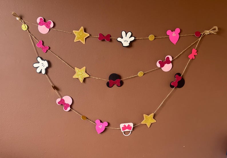 Puede incluir: Guirnalda decorativa con formas variadas: orejas de Mickey, lazos, estrellas y guantes. Las formas son rosas, negras, blancas y doradas con purpurina, ensartadas en cuerda. Para decorar fiestas.