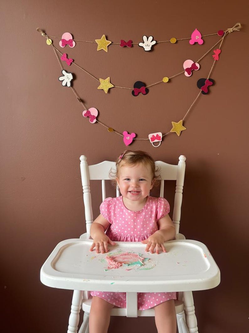 Puede incluir: Una ni&ntilde;a sonriente con un vestido rosa de lunares est&aacute; sentada en una trona blanca. Encima, una guirnalda de adornos con tem&aacute;tica de Minnie Mouse, incluyendo lazos, estrellas y guantes, cuelga de una cuerda.