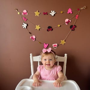 Puede incluir: Un beb&eacute; sonriente con un vestido y una diadema rosas est&aacute; sentado en una trona blanca. Una guirnalda decorativa con lazos rosas, estrellas y formas tem&aacute;ticas de Mickey Mouse cuelga sobre el beb&eacute;. El fondo es una pared marr&oacute;n lisa.