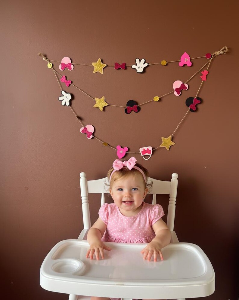 Puede incluir: Un beb&eacute; sonriente con un vestido y una diadema rosas est&aacute; sentado en una trona blanca. Una guirnalda decorativa con lazos rosas, estrellas y formas tem&aacute;ticas de Mickey Mouse cuelga sobre el beb&eacute;. El fondo es una pared marr&oacute;n lisa.
