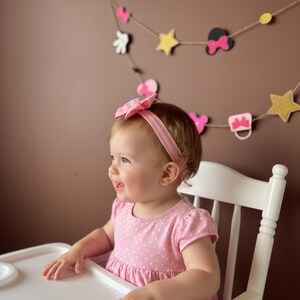 Puede incluir: Una ni&ntilde;a peque&ntilde;a con un vestido rosa de lunares y una diadema a juego est&aacute; sentada en una trona blanca. Una guirnalda decorativa con lazos, estrellas y siluetas de Mickey Mouse cuelga en el fondo.