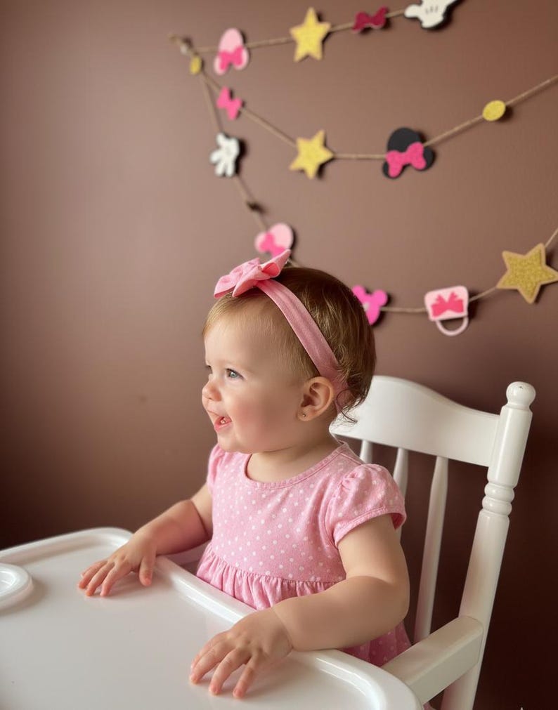 Puede incluir: Una ni&ntilde;a peque&ntilde;a con un vestido rosa de lunares y una diadema a juego est&aacute; sentada en una trona blanca. Una guirnalda decorativa con lazos, estrellas y siluetas de Mickey Mouse cuelga en el fondo.