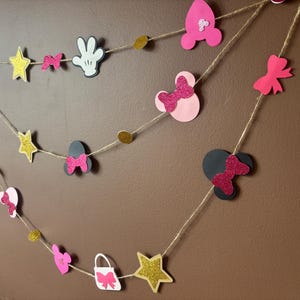 Puede incluir: Una pancarta decorativa con formas de estrella, lazos y siluetas de Mickey Mouse. Las formas son rosas, negras, blancas y doradas, ensartadas con hilo. Ideal para una fiesta o celebraci&oacute;n.