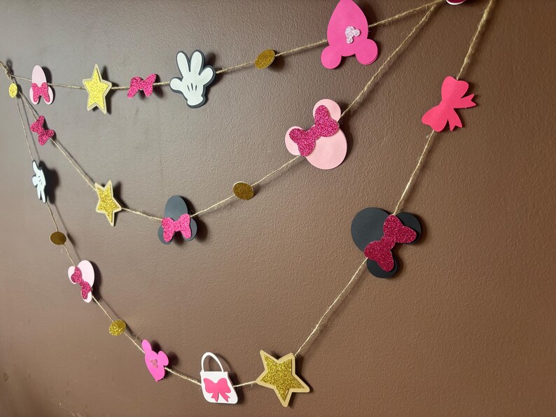 Puede incluir: Una pancarta decorativa con formas de estrella, lazos y siluetas de Mickey Mouse. Las formas son rosas, negras, blancas y doradas, ensartadas con hilo. Ideal para una fiesta o celebraci&oacute;n.