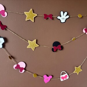 Puede incluir: Guirnalda decorativa con recortes de orejas de Mickey Mouse, lazos, estrellas y guantes en rosa, negro, blanco y purpurina dorada. Las formas est&aacute;n ensartadas en hilo, creando una decoraci&oacute;n festiva.