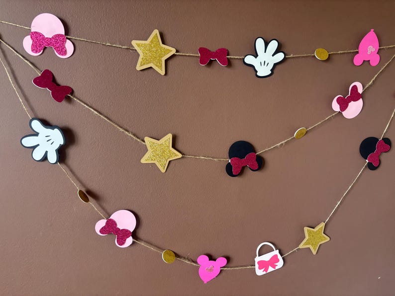 Puede incluir: Guirnalda decorativa con recortes de orejas de Mickey Mouse, lazos, estrellas y guantes en rosa, negro, blanco y purpurina dorada. Las formas est&aacute;n ensartadas en hilo, creando una decoraci&oacute;n festiva.
