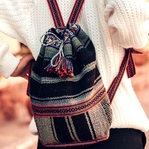 May include: A woven backpack with a drawstring closure and patterned straps. The bag features a black and gray striped design with red and white accents. A tassel hangs from the top.