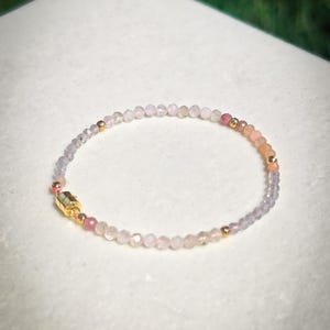 May include: A delicate beaded bracelet featuring small, faceted, translucent stones in shades of pale pink, lavender, and peach. The bracelet is accented with tiny, polished gold-tone metal beads and includes a cylindrical gold-tone magnetic clasp.