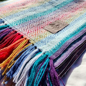 May include: A colorful woven blanket with a diamond pattern and a fringe of tassels in various colors. The blanket features a rainbow of colors, including red, yellow, blue, and purple. A small tag with the text "ALPACA SOFT" is attached to the blanket.
