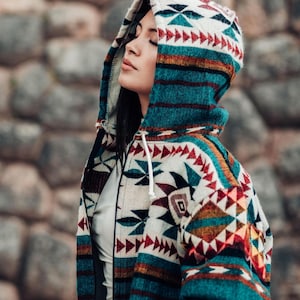 May include: A hooded jacket with a colorful geometric pattern in teal, red, white, and brown. The jacket has a zipper closure and a drawstring hood. The background is a stone wall.