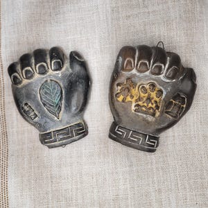 May include: Two fist-shaped wall decorations. One is dark grey with a leaf design, the other is brown with a gold relief. Both have a Greek key design at the base.