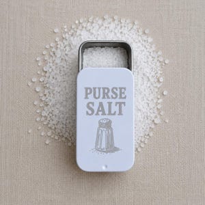 Pode incluir: Uma lata branca de sal de bolso com as palavras "PURSE SALT" e uma ilustração de um saleiro. A lata está aberta, revelando cristais de sal, e é rodeada por uma pilha de sal numa superfície de tom neutro.