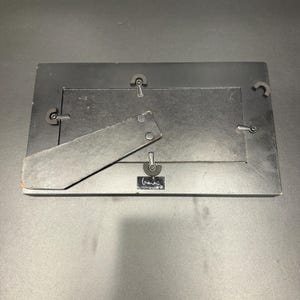 May include: A rectangular black picture frame shown from the back, featuring a hinged support stand, four metal swivel clips to secure the backing, and a small rectangular label with white text at the bottom center of the frame structure.
