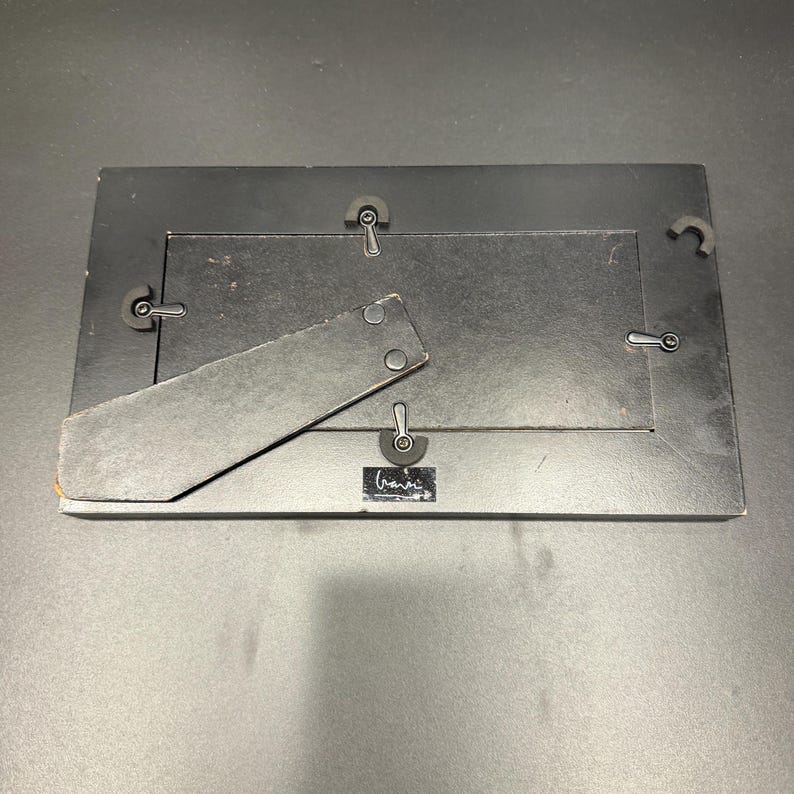 May include: A rectangular black picture frame shown from the back, featuring a hinged support stand, four metal swivel clips to secure the backing, and a small rectangular label with white text at the bottom center of the frame structure.