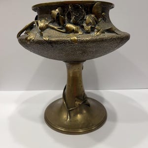 Puede incluir: Un cuenco de bronce sobre pedestal con superficie texturizada y relieves de hojas y enredaderas que envuelven la amplia cuenca superior y el tallo delgado. El metal tiene una pátina oscura y envejecida con reflejos pulidos en los detalles.