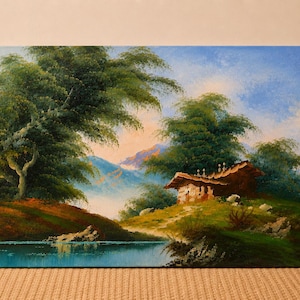 May include: Oil painting of a landscape featuring a small house nestled among green trees and rolling hills. A calm lake reflects the sky and surrounding foliage. The artwork is framed and ready to hang.
