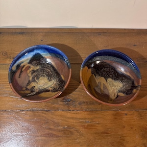 May include: Two handmade ceramic bowls with a unique, abstract design. The bowls feature a dark blue rim that fades into brown and gold tones in the center. The bowls are set on a wooden surface.