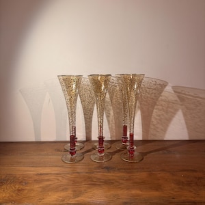 May include: Set of six champagne flutes with gold and black speckled design on the glass. Each flute has a red base and stem. The flutes are displayed on a wooden surface.