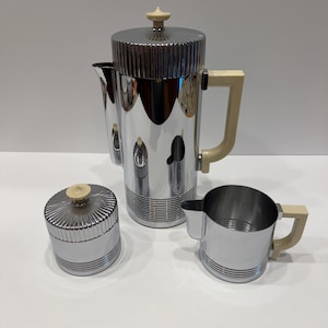 Puede incluir: Un juego de café de tres piezas que incluye una cafetera alta, un azucarero con tapa y una jarra para leche. Cada pieza está hecha de cromo pulido con una textura acanalada en la base y la tapa, complementada con asas y pomos de plástico color crema.