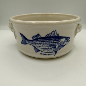May include: A round, off-white ceramic bowl with two side handles, featuring a blue line-art illustration of a fish on the front. Below the fish, the text "Striped Bass" is printed in blue. The bowl has a speckled, textured finish.