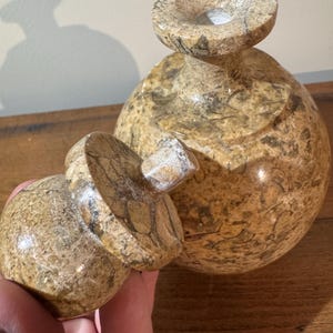 K&ouml;nnte beinhalten: Eine dekorative, handgeschnitzte Steinvase mit passendem Deckel. Die Vase hat eine runde Form mit einer weiten &Ouml;ffnung oben. Der Stein hat ein gesprenkeltes Muster aus Beige, Braun und Grau.