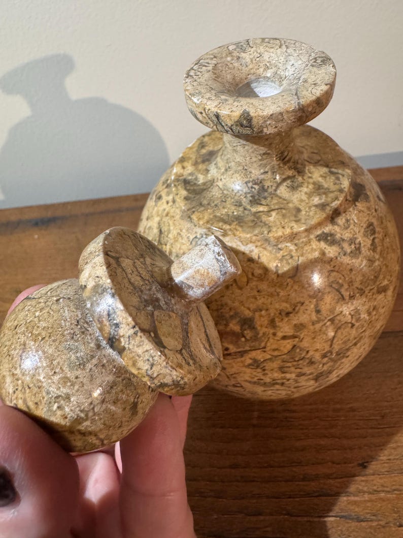 K&ouml;nnte beinhalten: Eine dekorative, handgeschnitzte Steinvase mit passendem Deckel. Die Vase hat eine runde Form mit einer weiten &Ouml;ffnung oben. Der Stein hat ein gesprenkeltes Muster aus Beige, Braun und Grau.