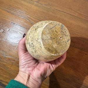 K&ouml;nnte beinhalten: Eine runde, beige Steinschale mit einem geschnitzten Blumenmuster. Die Schale wird in einer Hand gehalten, wodurch ihre Form und Textur zur Geltung kommen. Das Innere der Schale ist sichtbar und zeigt ihre Tiefe und die nat&uuml;rlichen Variationen des Steins.