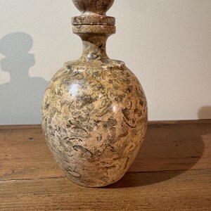 K&ouml;nnte beinhalten: Eine dekorative Flasche mit rundem K&ouml;rper und einem Verschluss. Die Flasche besteht aus einem gesprenkelten, hellbraunen Material mit dunkleren braunen und gr&uuml;nen Markierungen. Der Verschluss hat eine Kugelform. Die Flasche steht auf einer Holzoberfl&auml;che.
