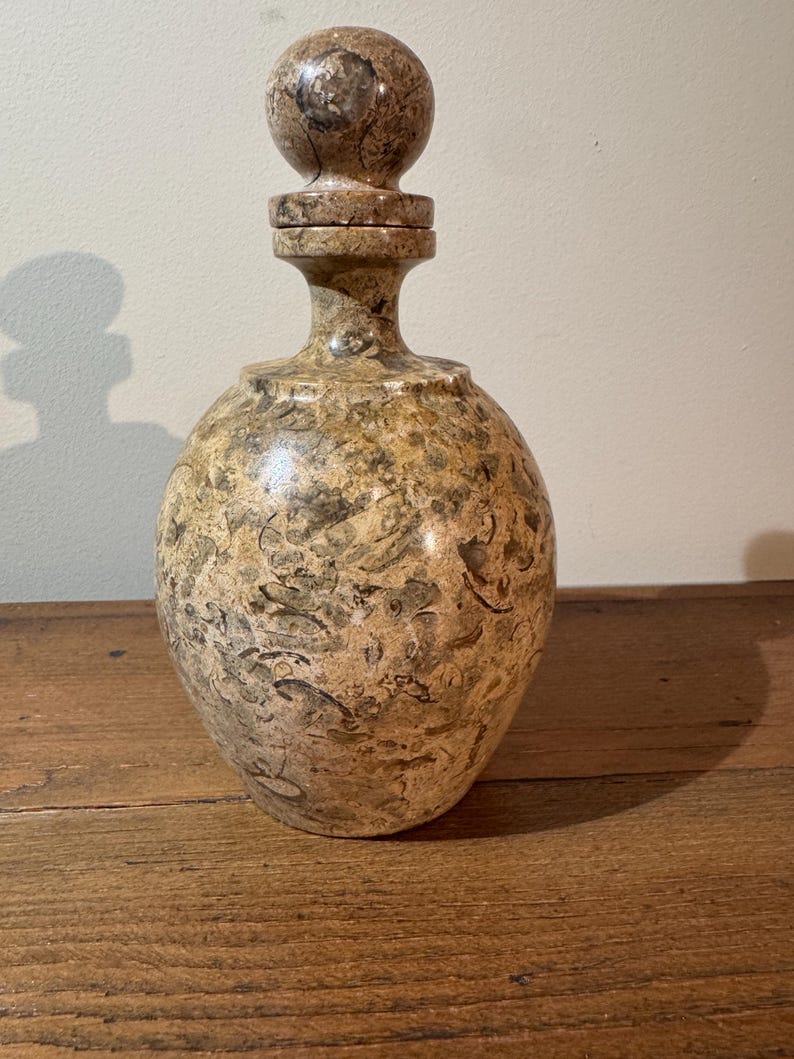 K&ouml;nnte beinhalten: Eine dekorative Flasche mit rundem K&ouml;rper und einem Verschluss. Die Flasche besteht aus einem gesprenkelten, hellbraunen Material mit dunkleren braunen und gr&uuml;nen Markierungen. Der Verschluss hat eine Kugelform. Die Flasche steht auf einer Holzoberfl&auml;che.