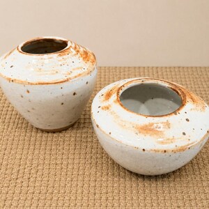 May include: Two handmade ceramic vases. The vases are white with brown speckles and a brown rim. One vase is round, and the other is oval. The vases are on a tan surface.