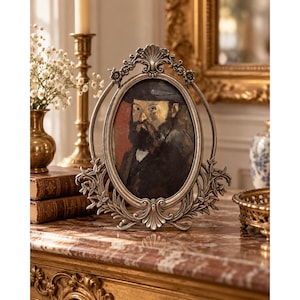 May include: An oval silver-toned metal picture frame with ornate scrollwork and floral detailing at the top and bottom. The frame holds a portrait painting of a bearded person wearing a dark hat against a red and dark background.
