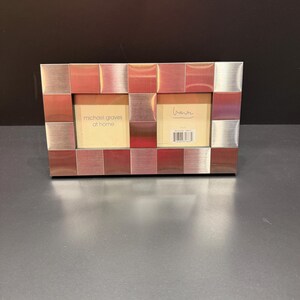 May include: A rectangular double photo frame featuring a grid of alternating brushed silver and copper-toned metal squares. The frame contains two cream-colored inserts, one with the text 'michael graves at home' and the other with a signature and barcode.